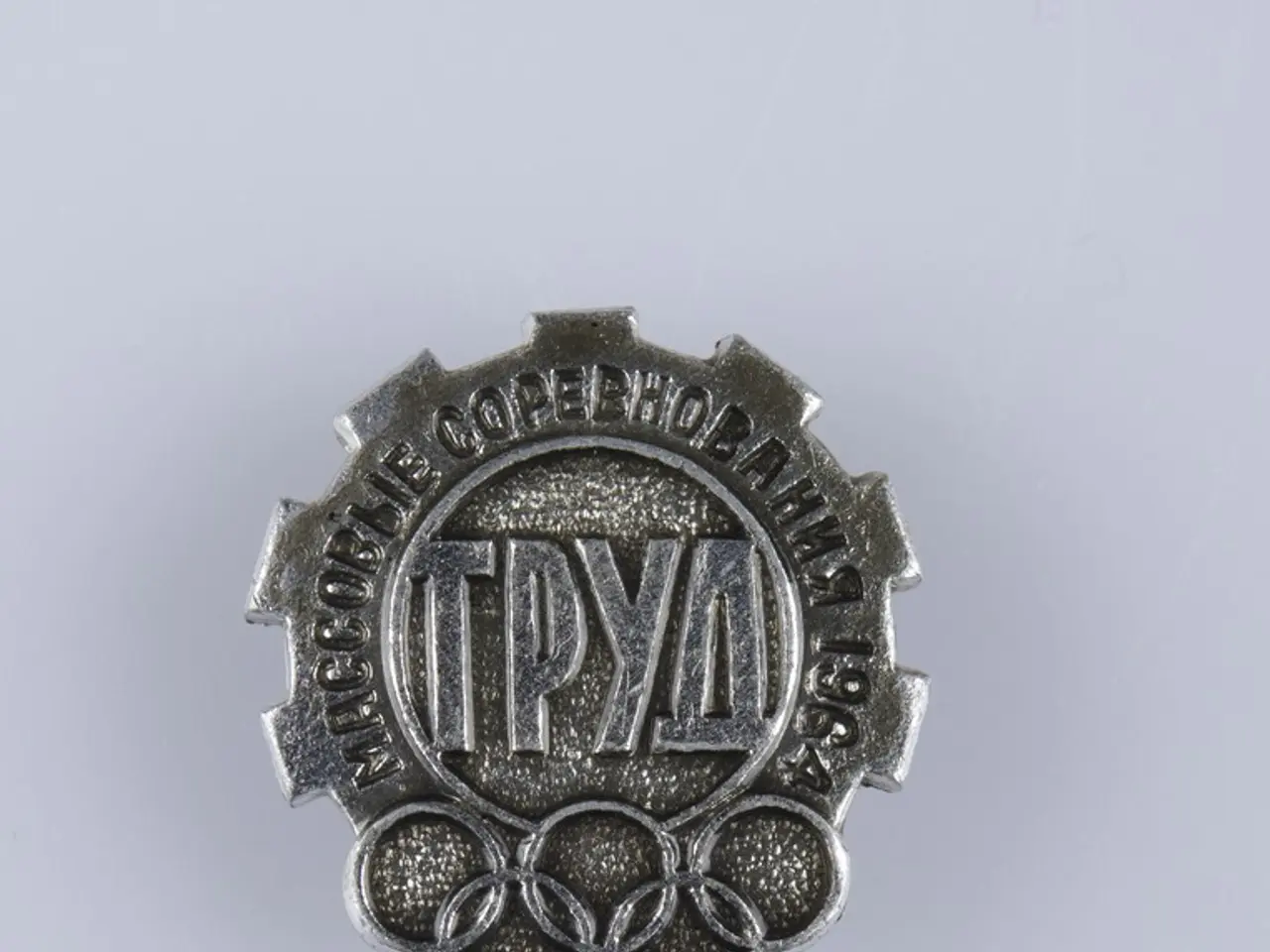 The image shows a silver badge with the words "Olympic Games" written on it against a white...