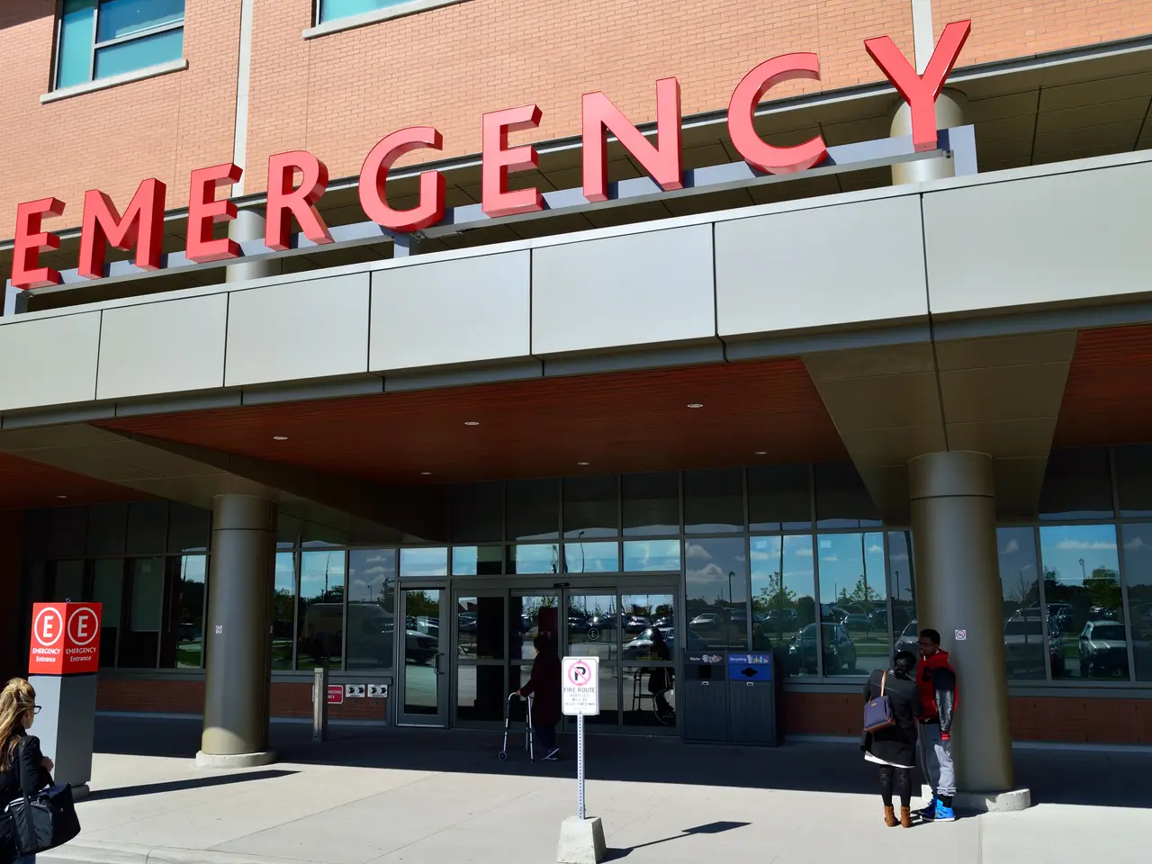 The image shows a group of people walking in front of an emergency building with glass walls and...