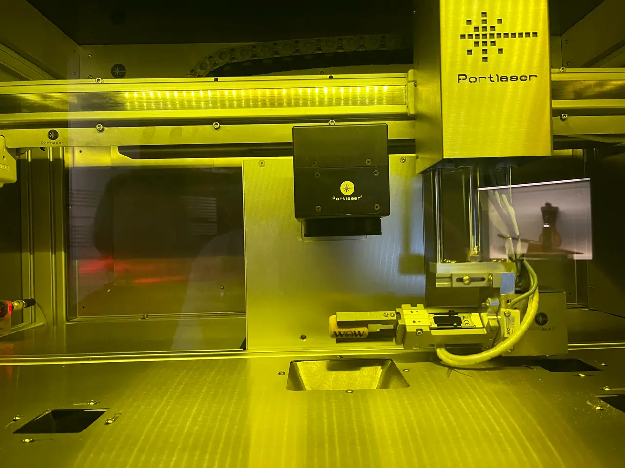 The image shows a 3D printer sitting on top of a table, with a screen in the background. The...
