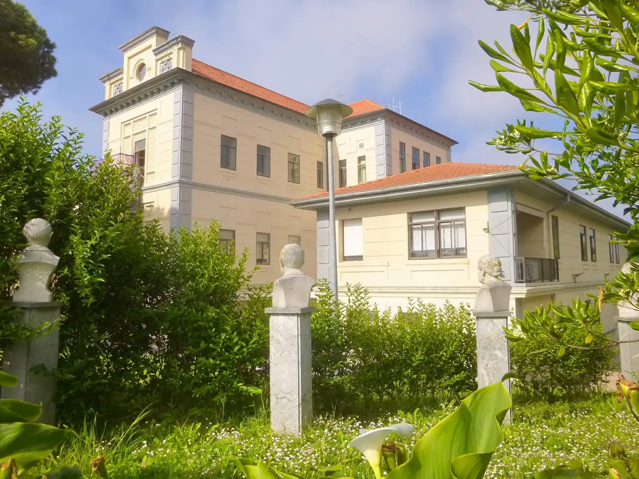 The image shows a large white building with a red roof in the middle of a lush green field,...