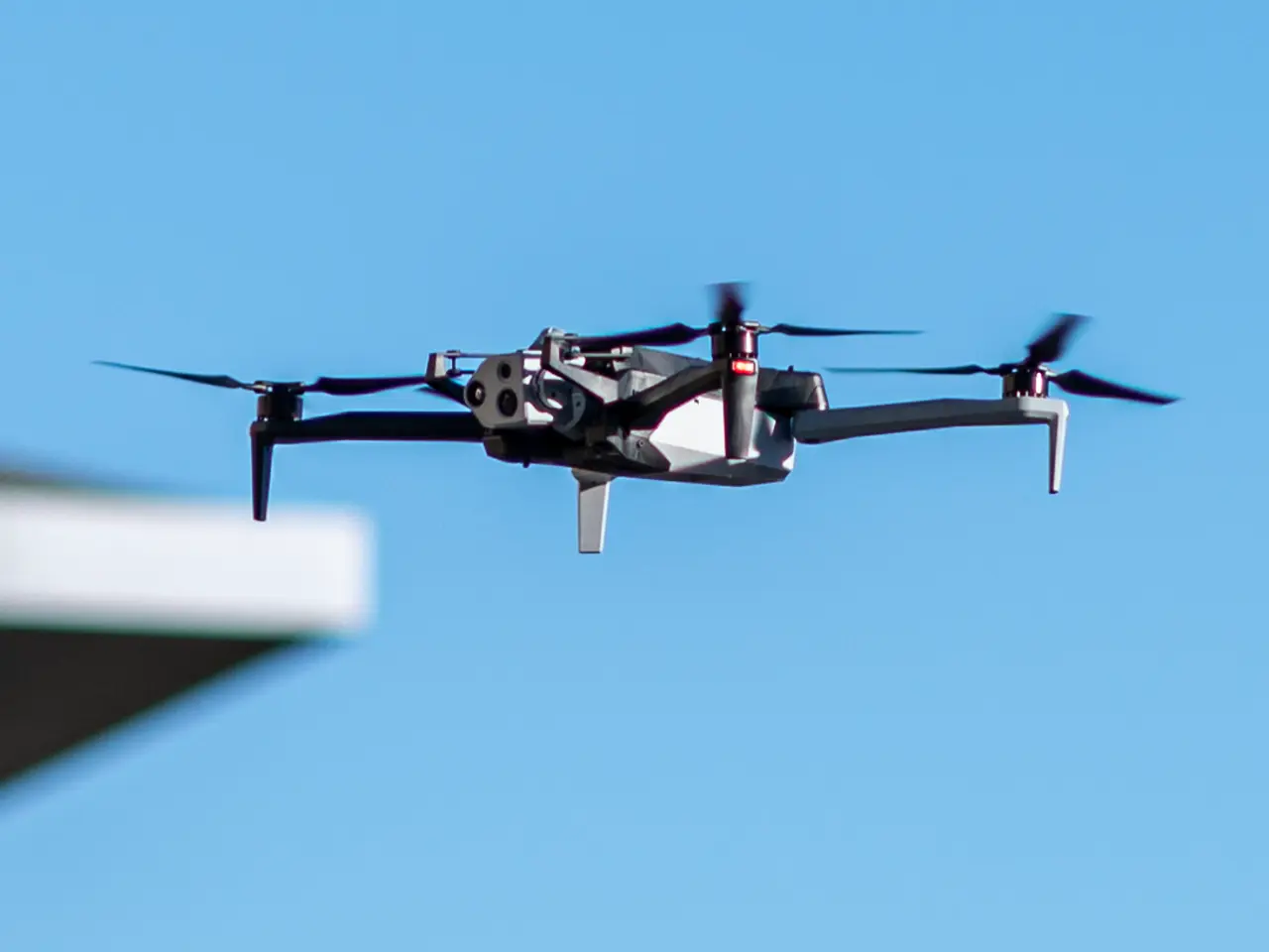 The image shows a drone flying in the air with a building on the left side and a clear blue sky in...
