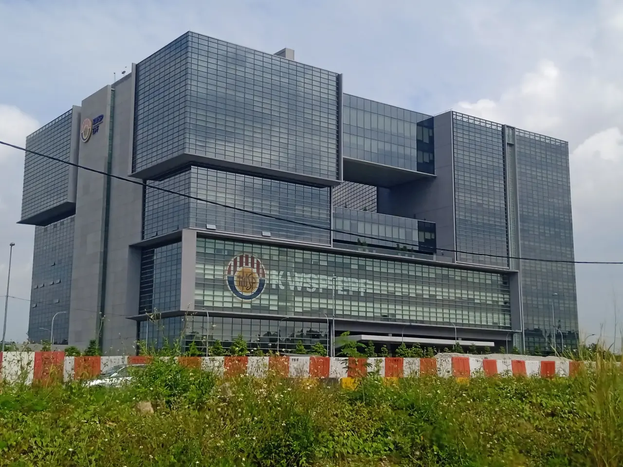 The image shows a large building with a lot of windows on the side of it, surrounded by plants, a...