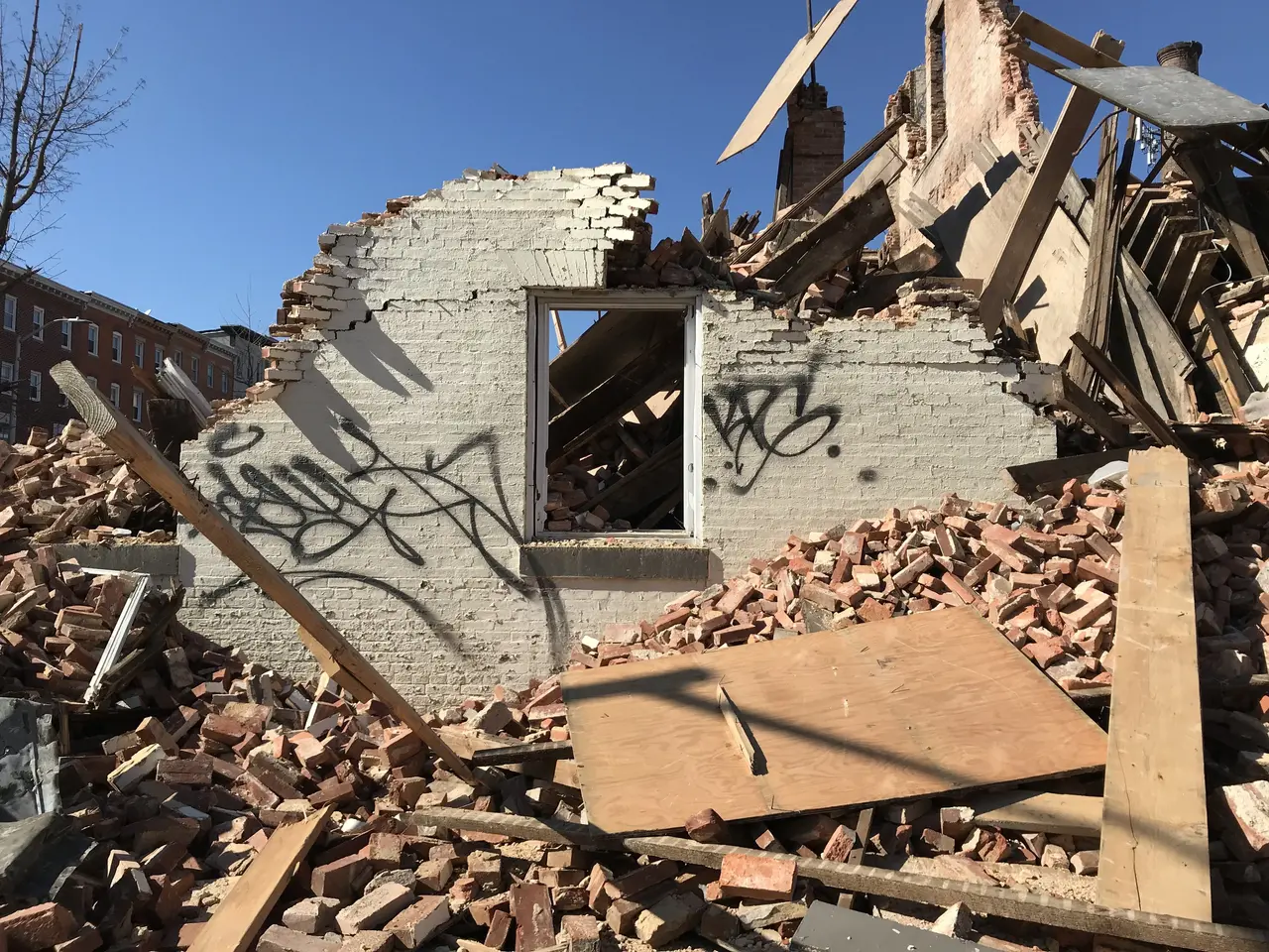 The image shows a demolished building with graffiti on the side of it, surrounded by bricks, wooden...