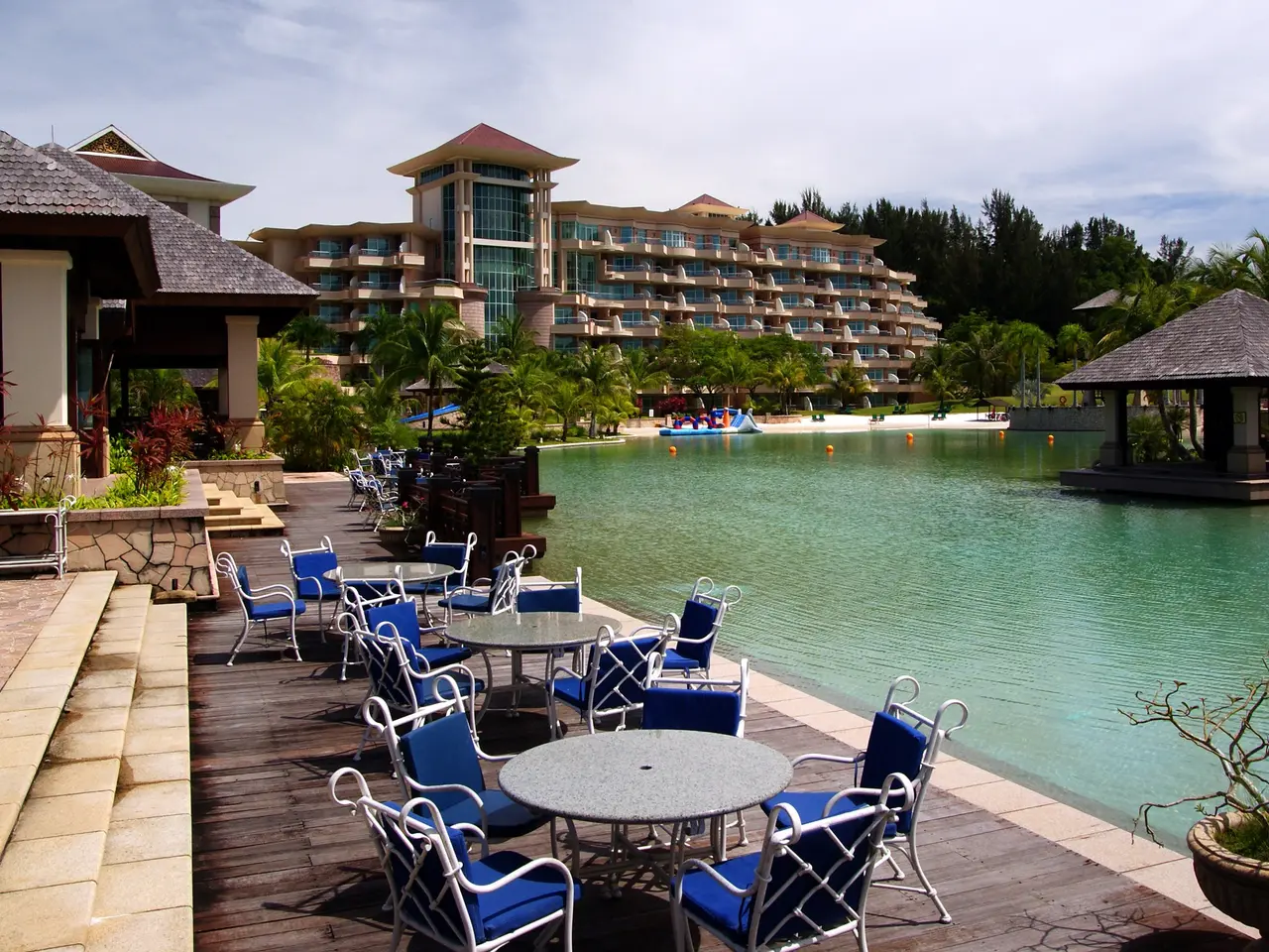 The image shows a resort with tables and chairs near a large body of water, surrounded by lush...