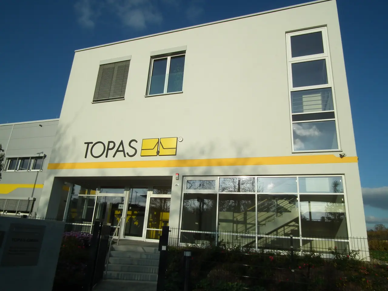 The image shows a building with glass windows and doors, steps with railings, plants with flowers,...