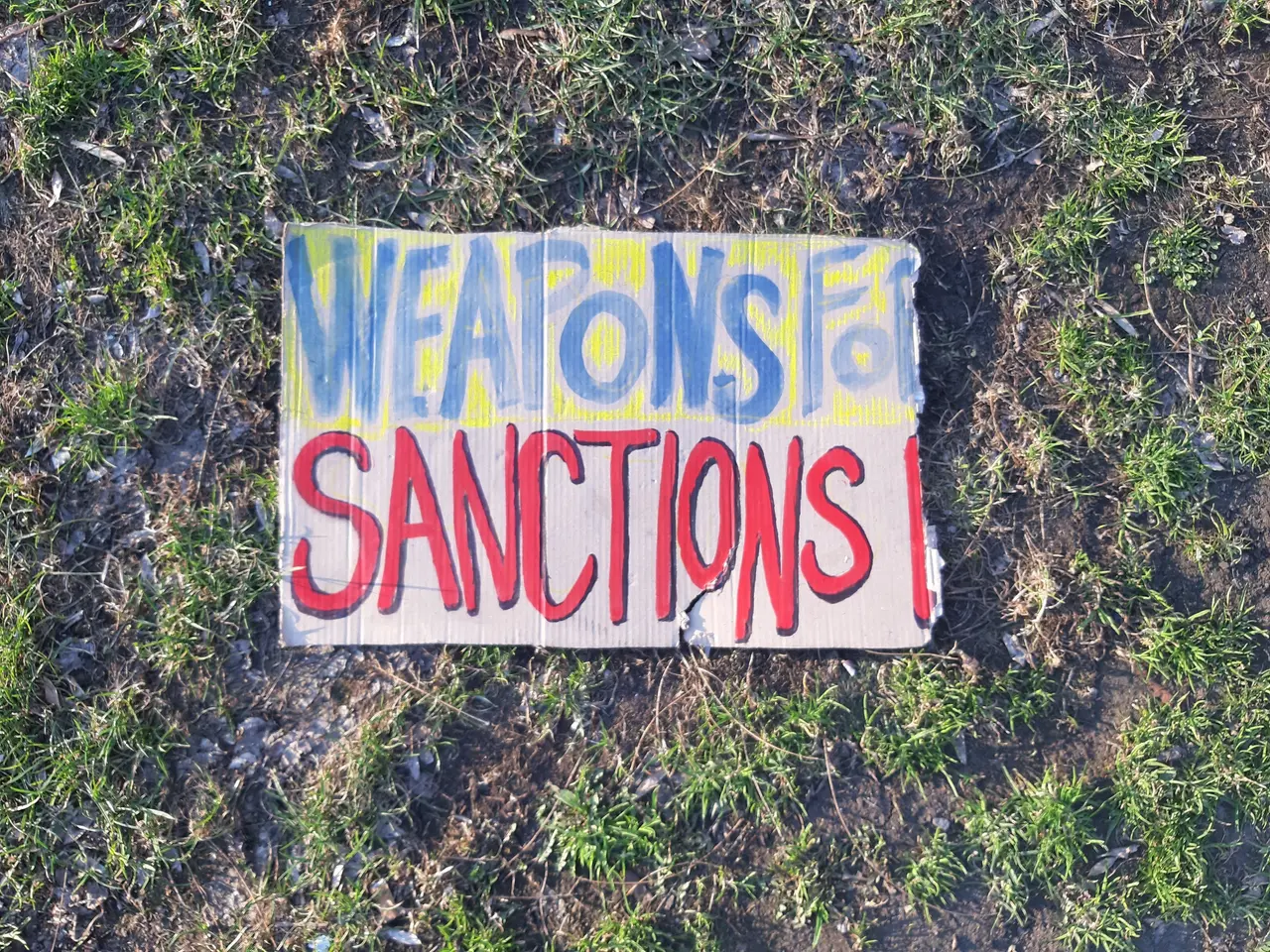 The image shows a sign on the ground that reads "Weapons for Sanctions" in bold black lettering...
