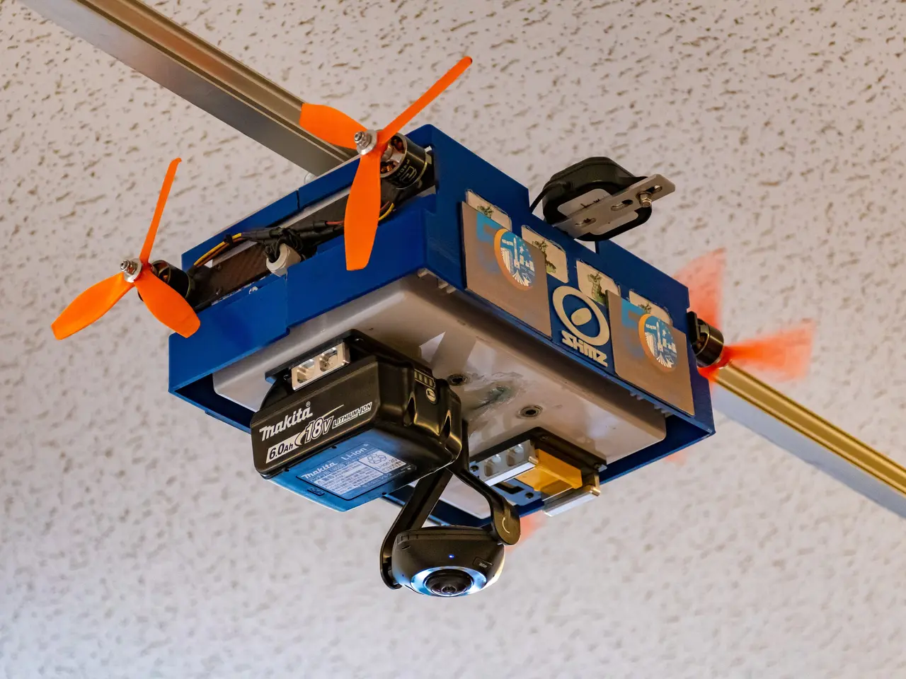 The image shows a small blue drone with orange propellers flying in the air, attached to a metal...