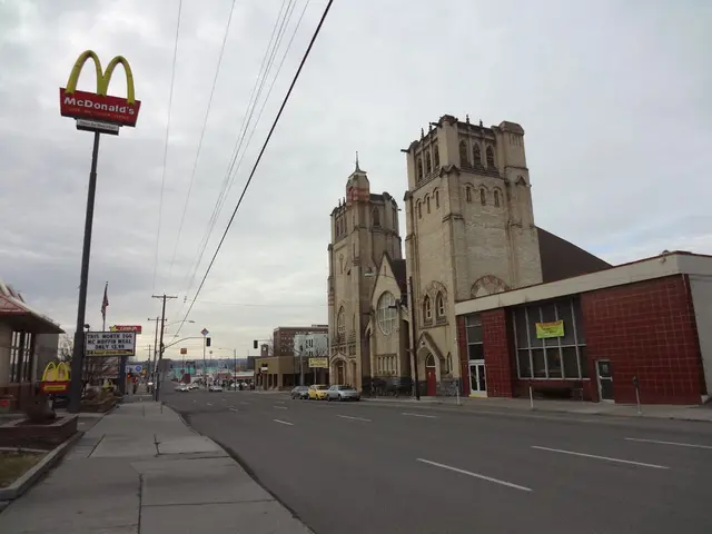 In this picture there are buildings on the right side of the image, there are poles and cars in the...