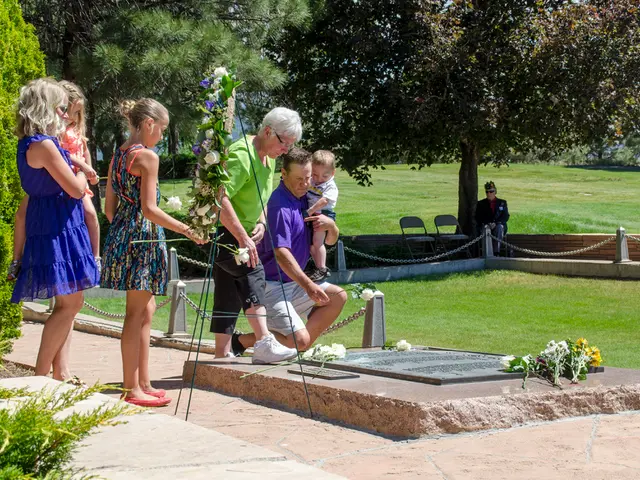 In the picture I can see group of persons standing, there is a person crouching near the gravestone...