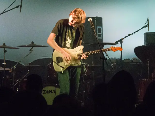 In this image there is a man standing and playing a guitar near a microphone and at back ground...