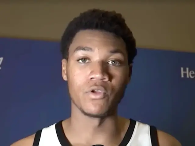 The image shows a man in a white tank top holding a microphone, with a banner in the background...