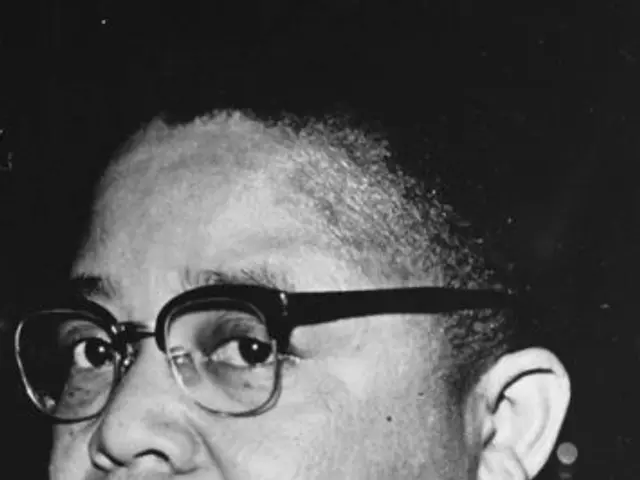 The image shows a black and white photo of Martin Luther King Jr. wearing a suit and spectacles,...