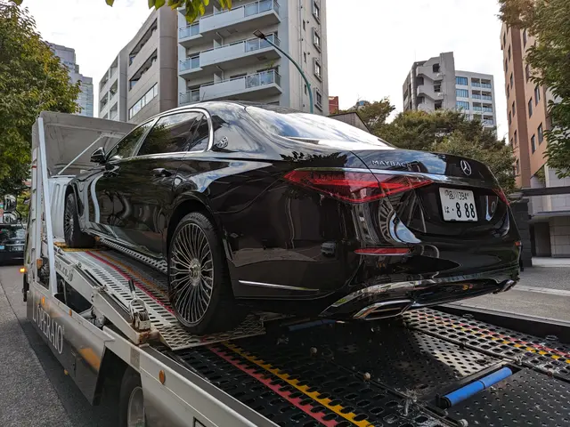 The image shows a black car being towed by a tow truck on a city street, surrounded by buildings,...