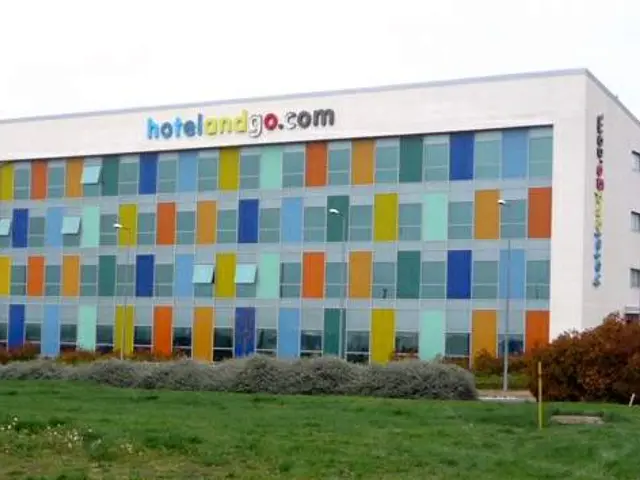 The image shows a large building with a lot of windows on the side of it, surrounded by plants,...