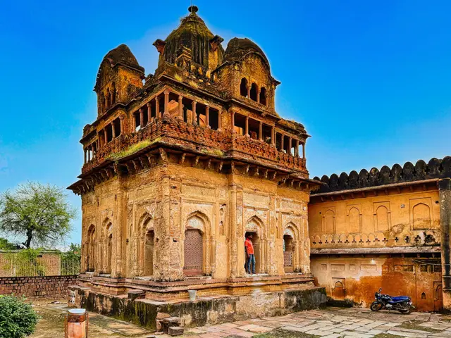 The image shows an old building with a man standing in front of it, surrounded by a wall, trees,...