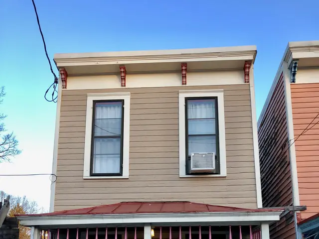 The image shows a two-story house with a fence in front of it, surrounded by plants, trees, and...