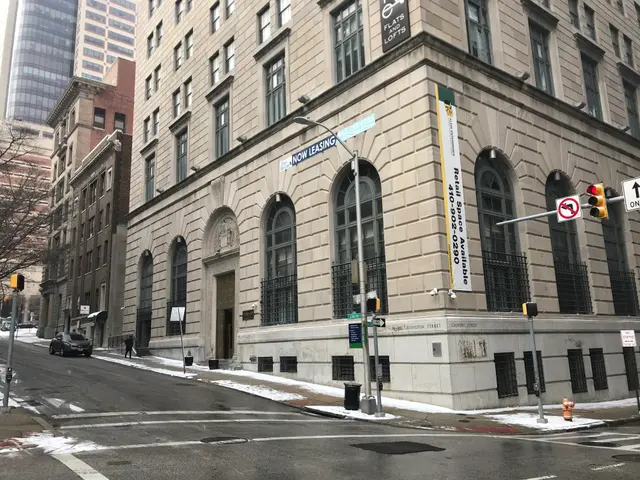 The image shows a large building on the corner of a city street, with vehicles driving by, light...