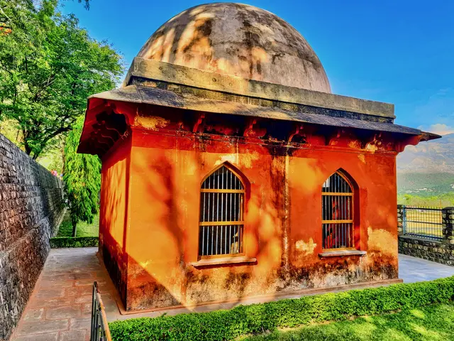 The image shows a small building with a dome on top of it, surrounded by a wall and a railing, with...