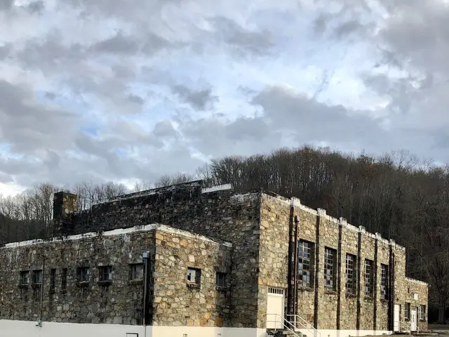 The image shows an old stone building sitting on the side of a road, surrounded by grass and trees,...