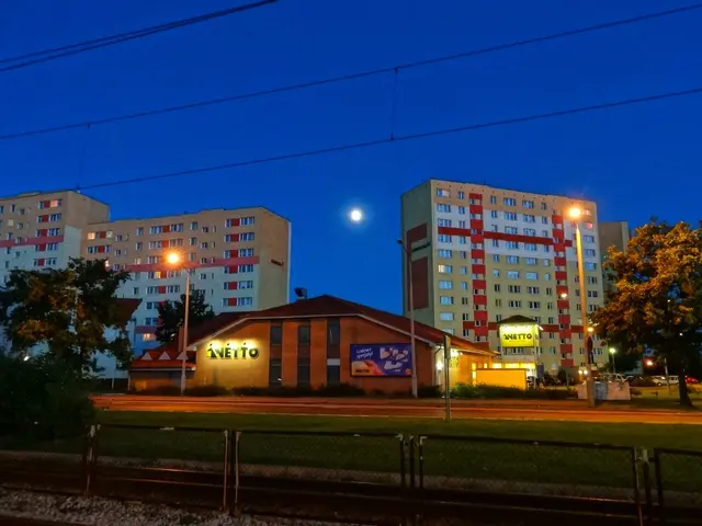 The image shows a city street at night with a full moon in the sky. We can see buildings with...