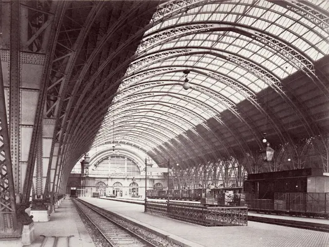 The image shows an old black and white photo of a train station with railway tracks, platforms,...