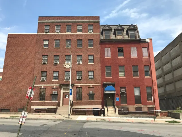 The image shows a large brick building on the corner of a street, with windows, doors, steps,...