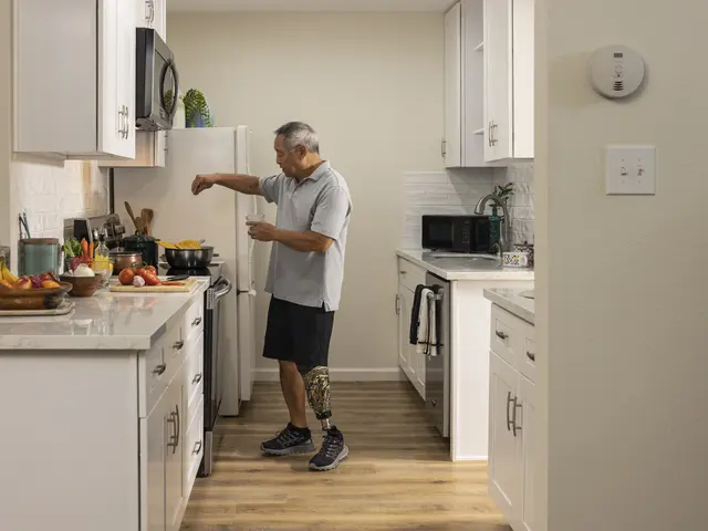 The image shows a man standing in a kitchen with white cabinets, holding a glass in his hand. On...