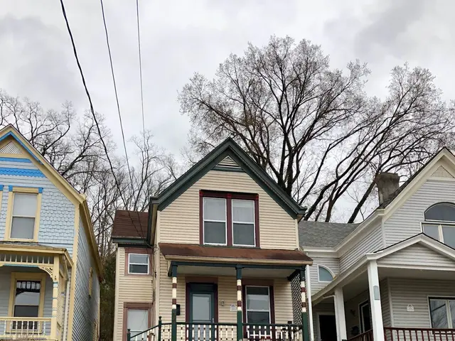 The image shows a black car parked in front of a row of multi-family homes for sale in the Bronx...