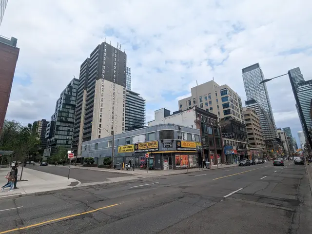 The image shows a city street with tall buildings in the background, vehicles on the road, people...