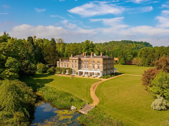The image shows an aerial view of a large house situated in the middle of a lush green field,...