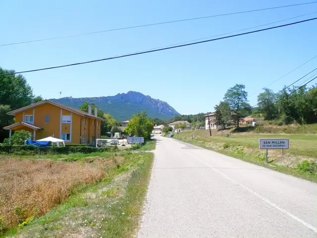 The image shows a road with a sign on the side of it, surrounded by grass, plants, trees, and...