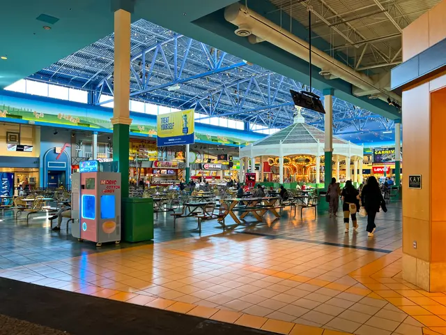The image shows the inside of a shopping mall with people walking around. There are tables and...